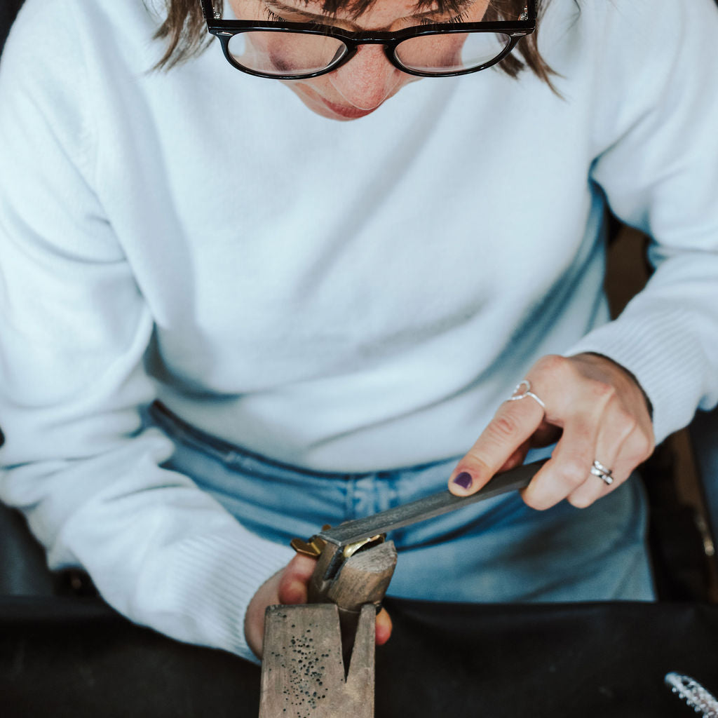 A close-up lifestyle shot of a jeweler wearing black-rimmed glasses and a white sweater, using calipers to measure a piece of jewelry held in a wooden stand on a workbench.