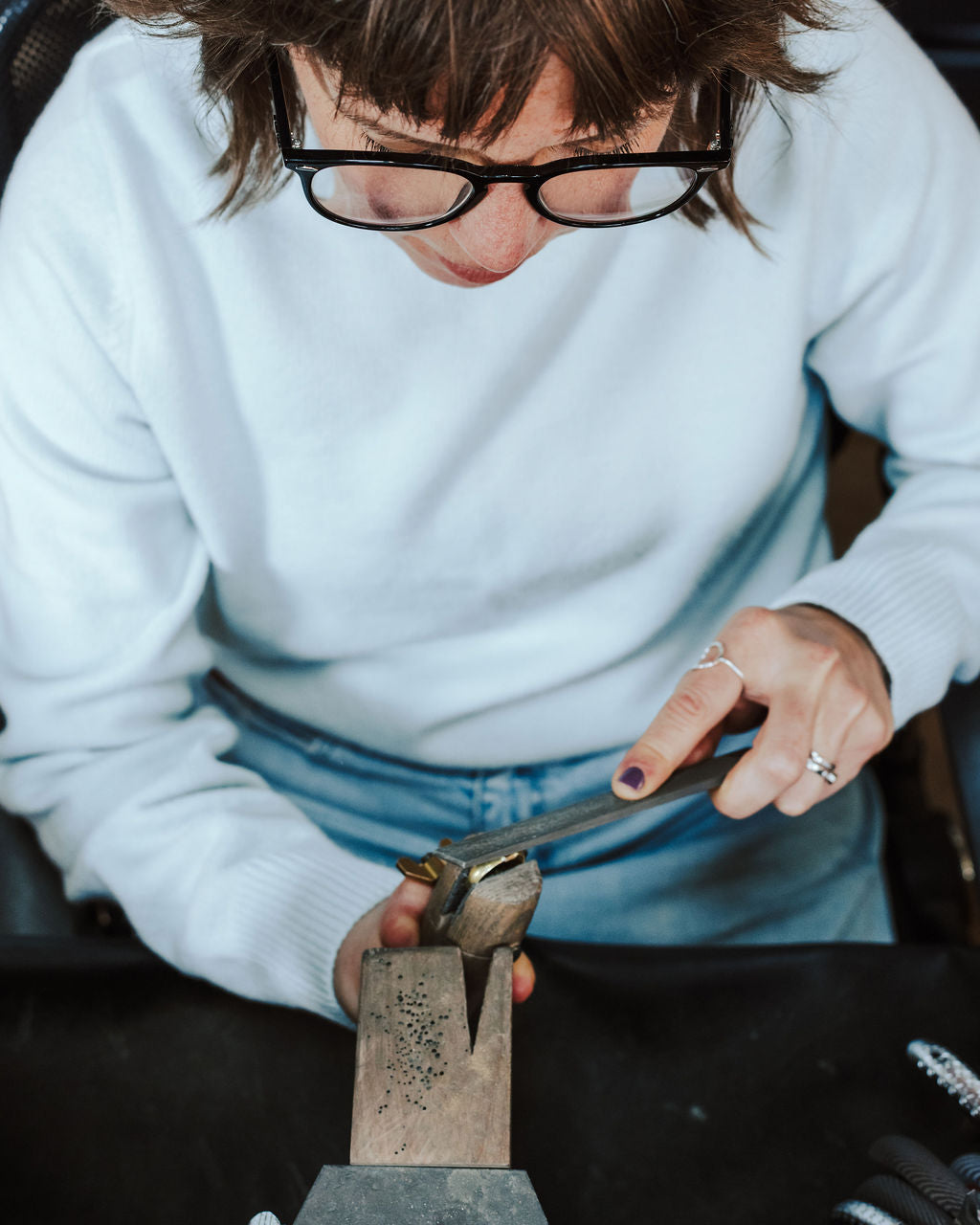 A close-up lifestyle shot of a jeweler wearing black-rimmed glasses and a white sweater, using calipers to measure a piece of jewelry held in a wooden stand on a workbench.