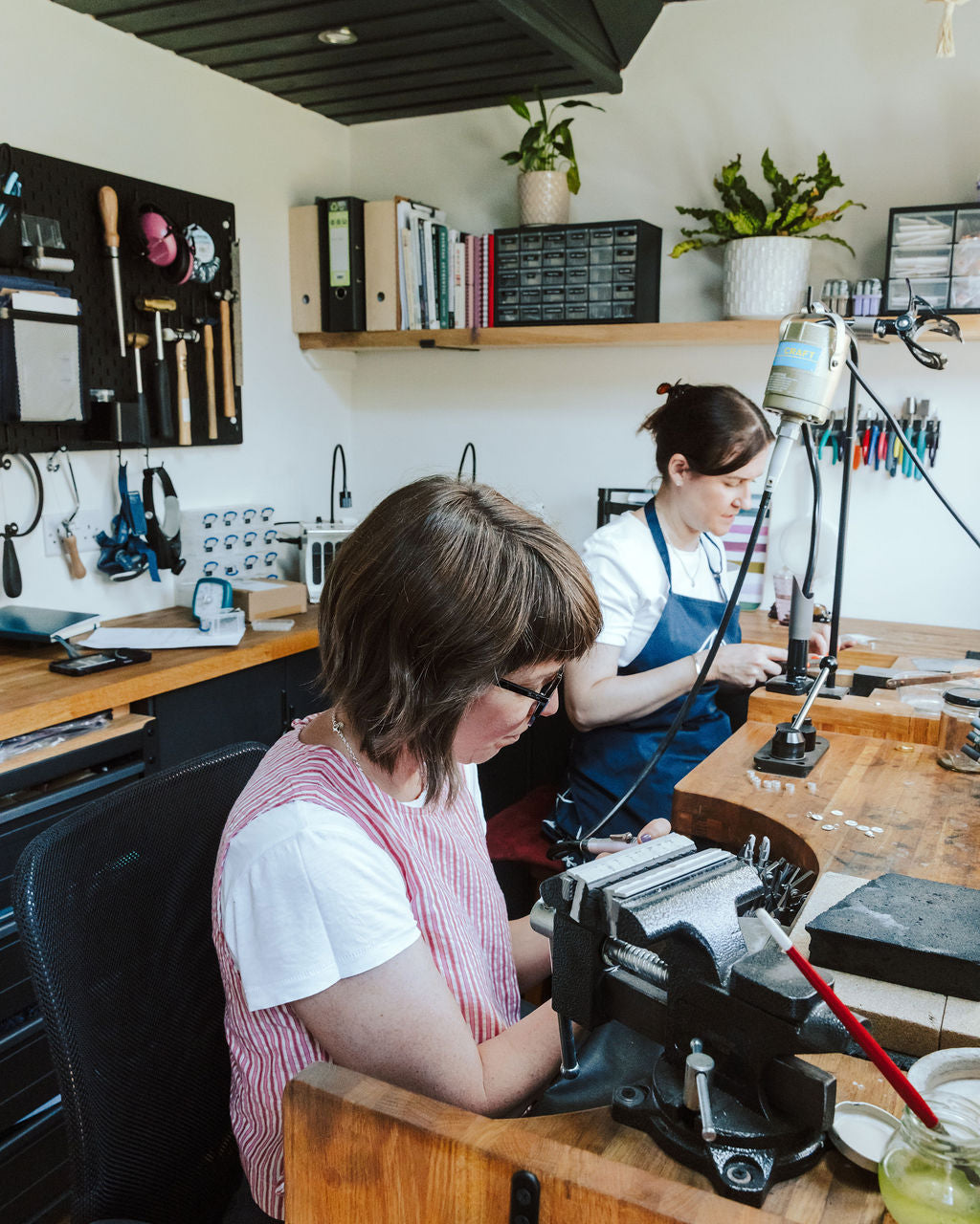 Two women working at a wooden craft desk in a workshop, one in the foreground using a black bench grinder and another in the background at her own station. The room has white walls, wooden shelves with plants and binders, and a dark ceiling.