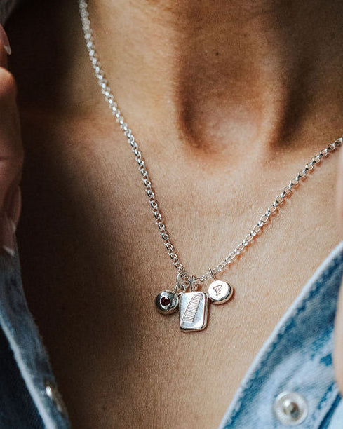 Woman in a denim shirt wearing a necklace featuring three charms, one birthstone, one feather and one initial.