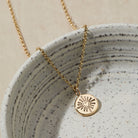 Gold necklace with a heart and sun design pendant on a speckled ceramic dish.