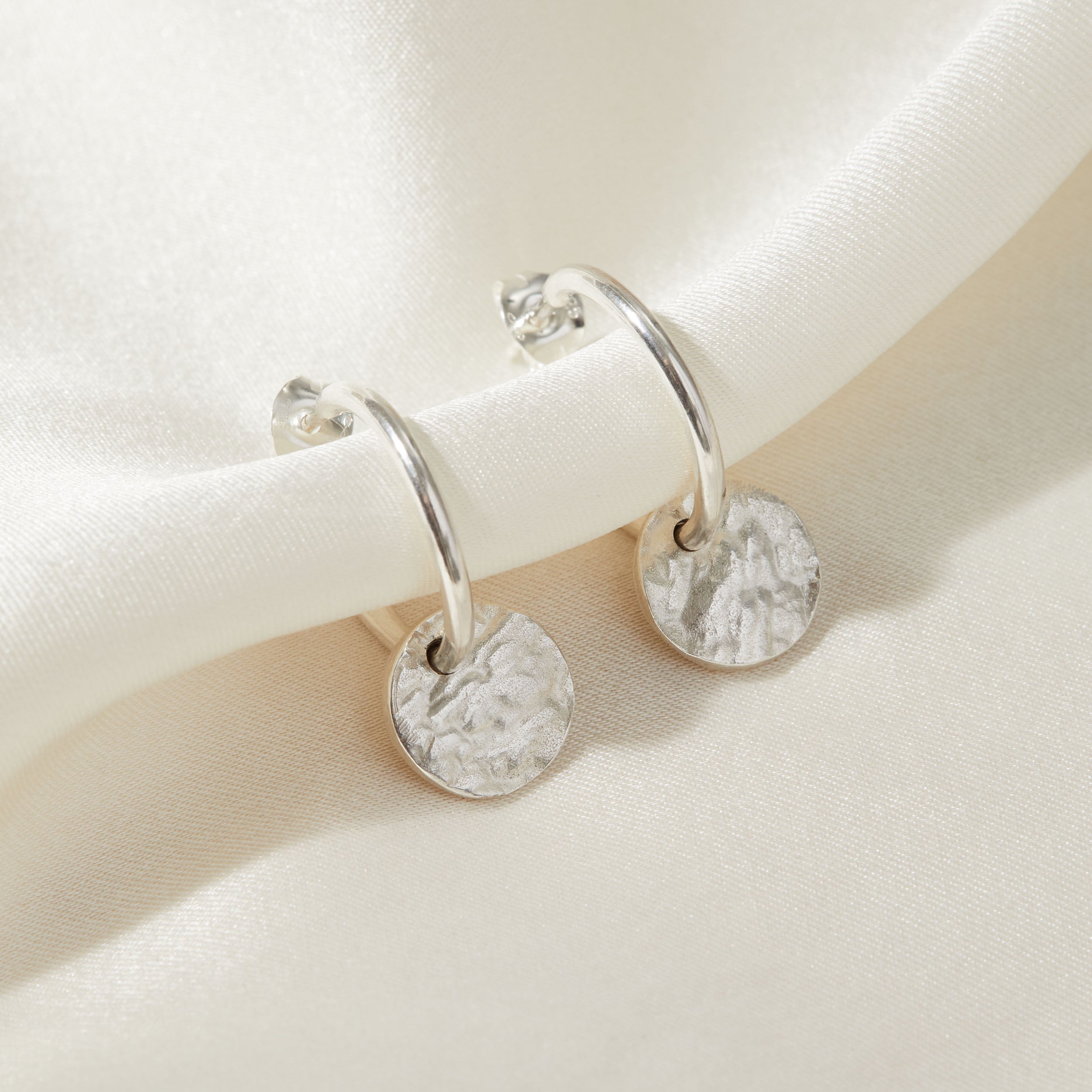 Sterling silver hoop earrings with bark textured charms on a beige background.