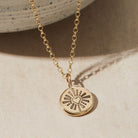 Gold necklace with a heart and sunburst pendant on a beige background