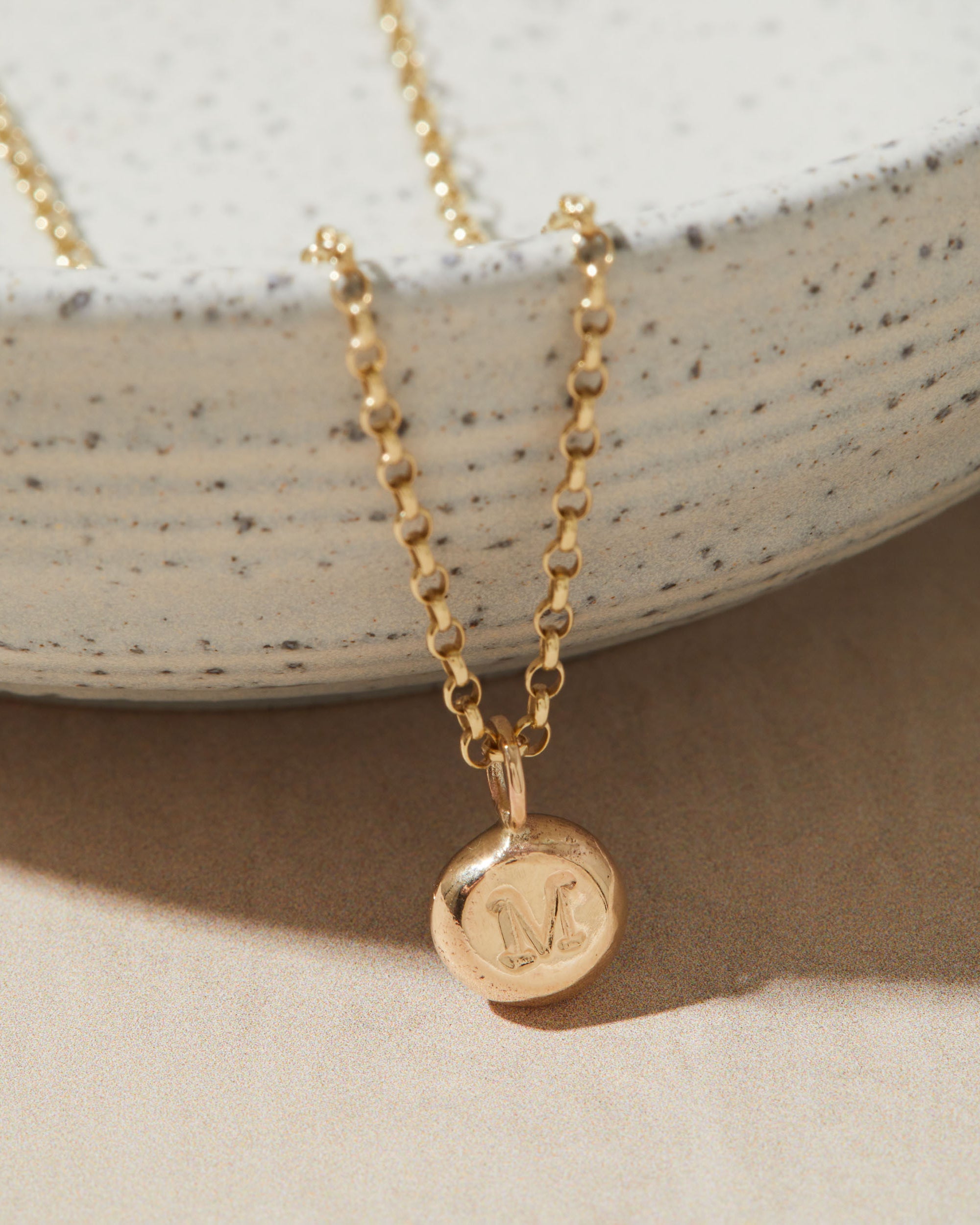 Gold necklace with a round pendant featuring the letter 'M' on a neutral background