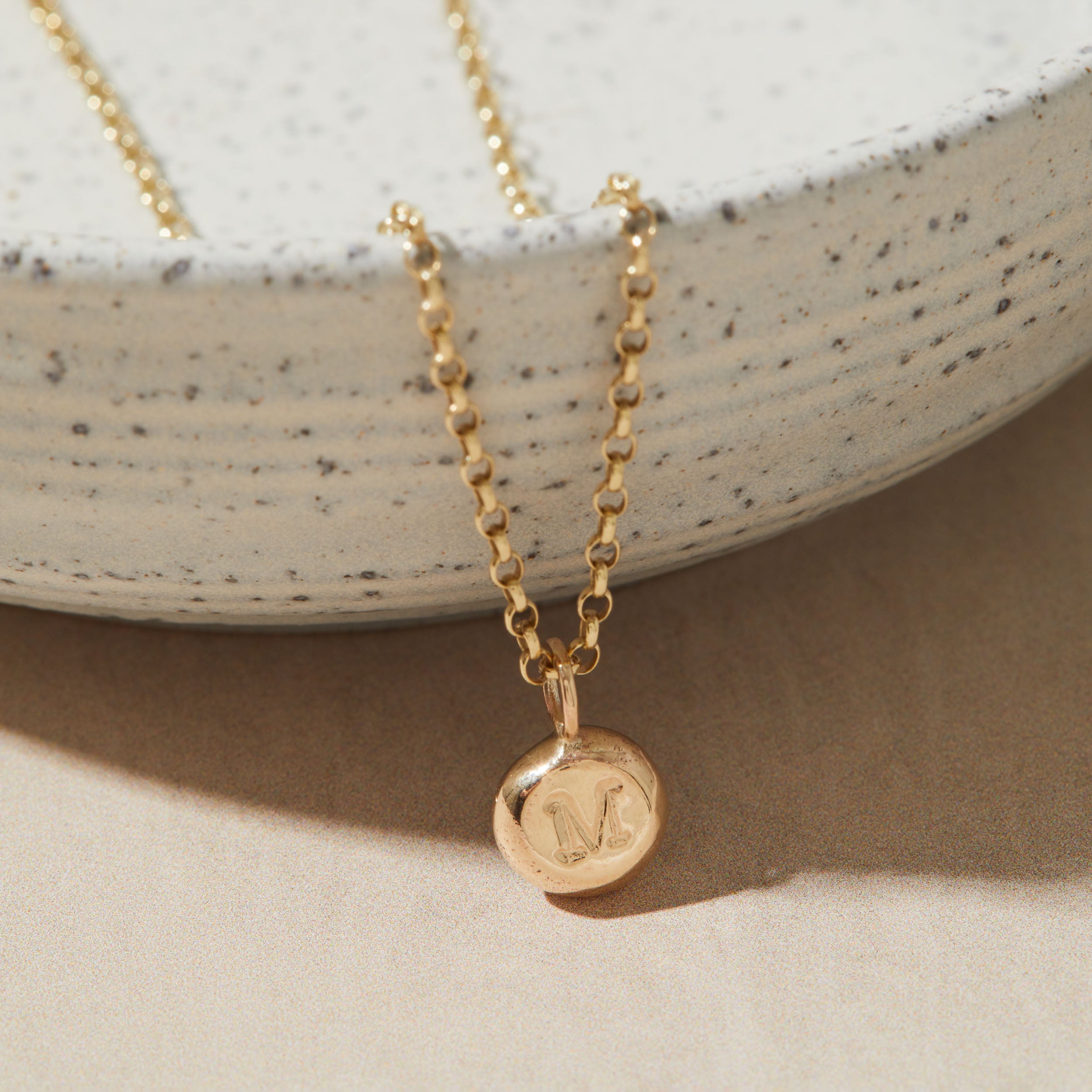 Gold necklace with a round pendant featuring the letter 'M' on a neutral background