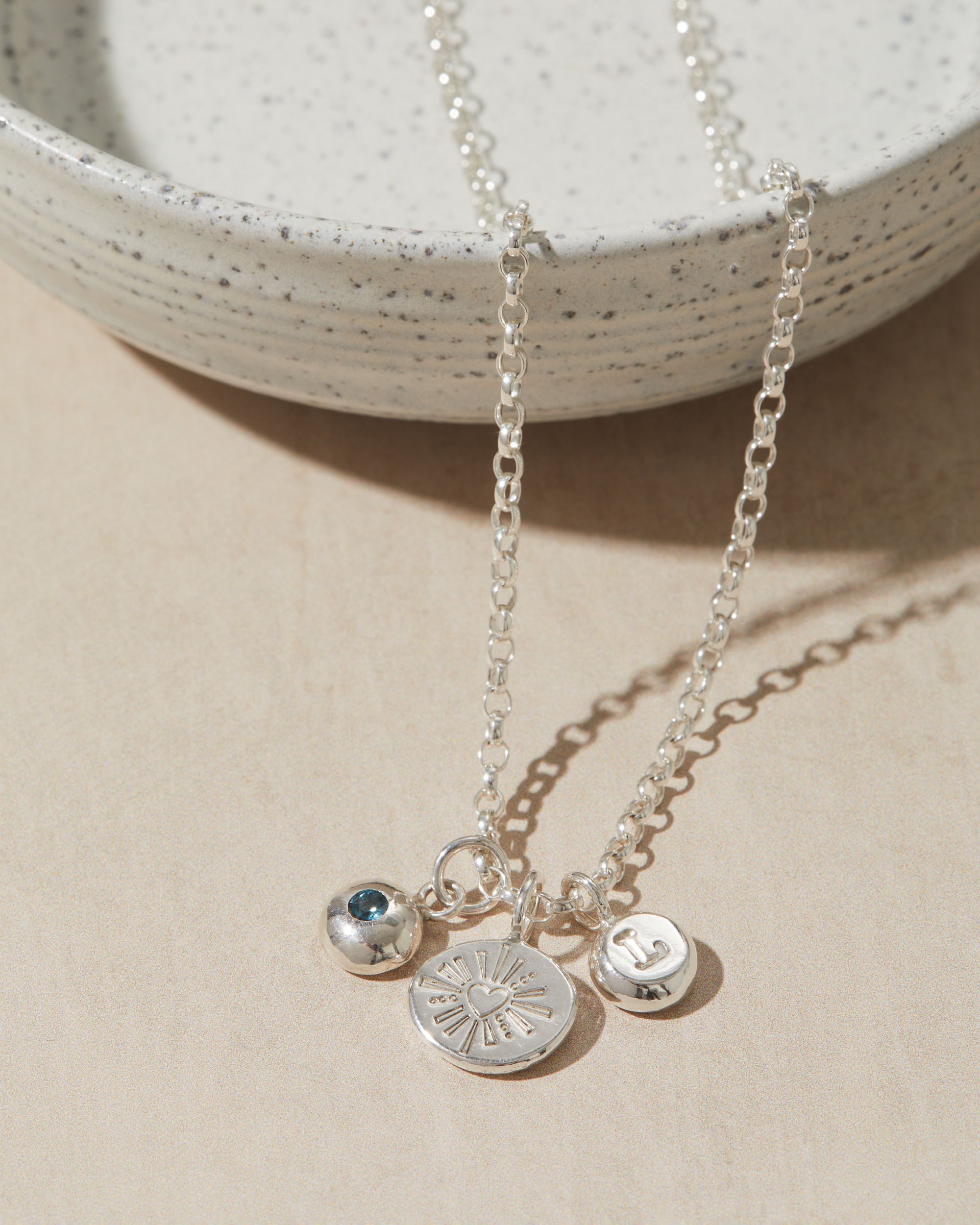 Silver necklace with heart charm, initial charm and birthstone charm on neutral surface.