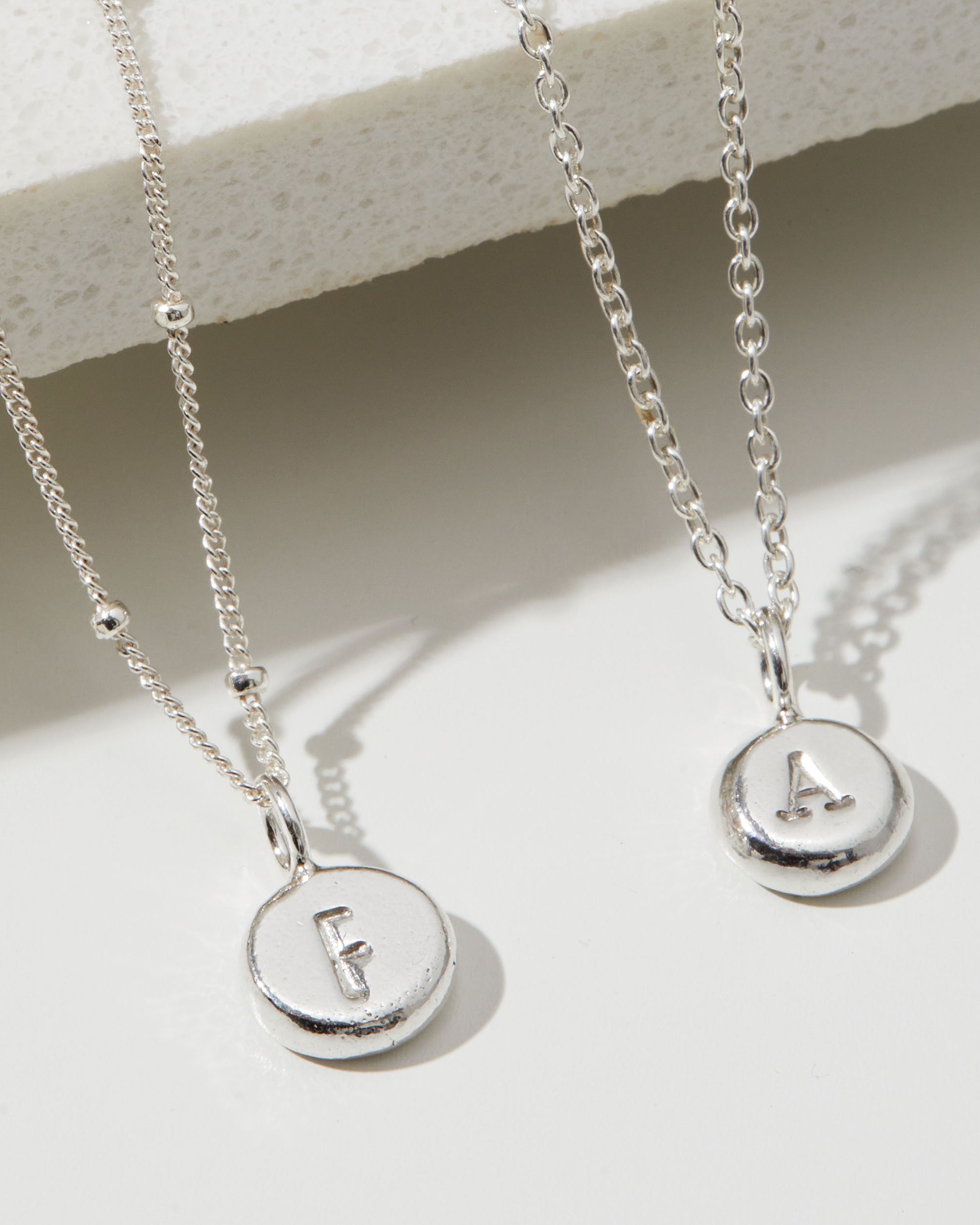 Two silver necklaces with round pendants displaying letters 'F' and 'A' on a light background.