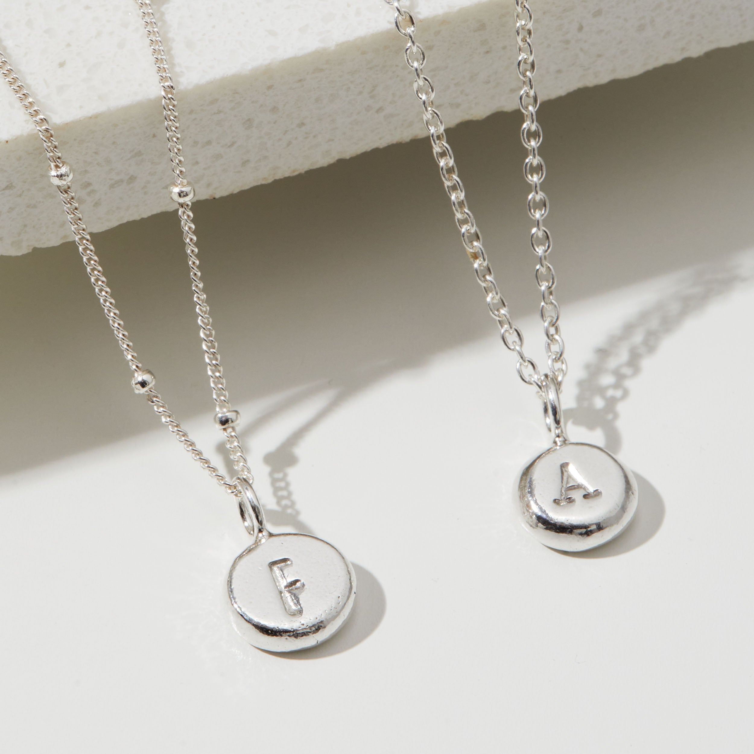 Two silver necklaces with round pendants displaying letters 'F' and 'A' on a light background.