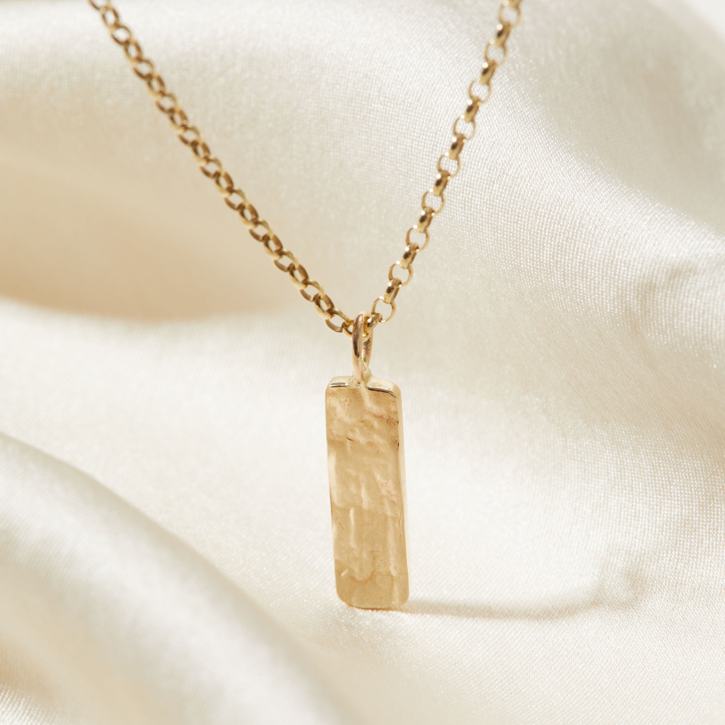 Gold necklace with a rectangular bark textured pendant on a beige fabric background