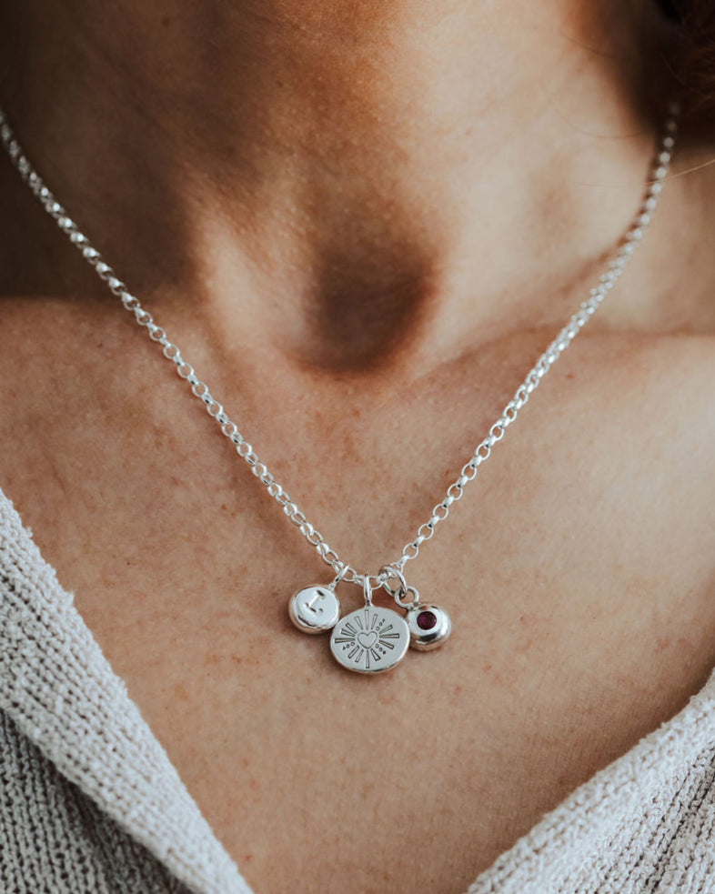 Silver necklace with heart, initial and birthstone pendants on a person wearing a light gray sweater.