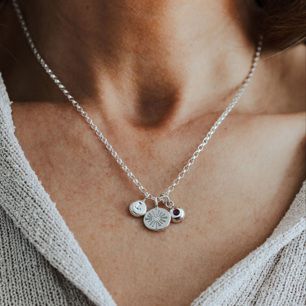 Silver necklace with heart, initial and birthstone pendants on a person wearing a light gray sweater.