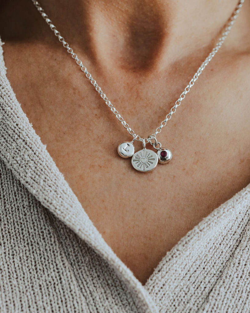 Silver necklace with initial, heart and birthstone pendants on a person wearing a beige sweater.