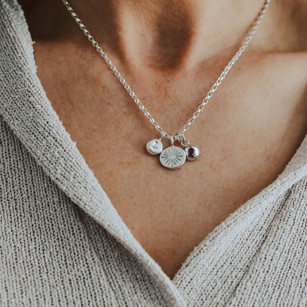 Silver necklace with initial, heart and birthstone pendants on a person wearing a beige sweater.