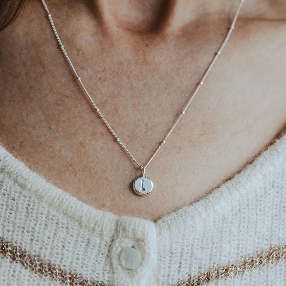 Necklace with a small initial pebble pendant on a pebble chain worn by a person in a white and brown striped sweater.
