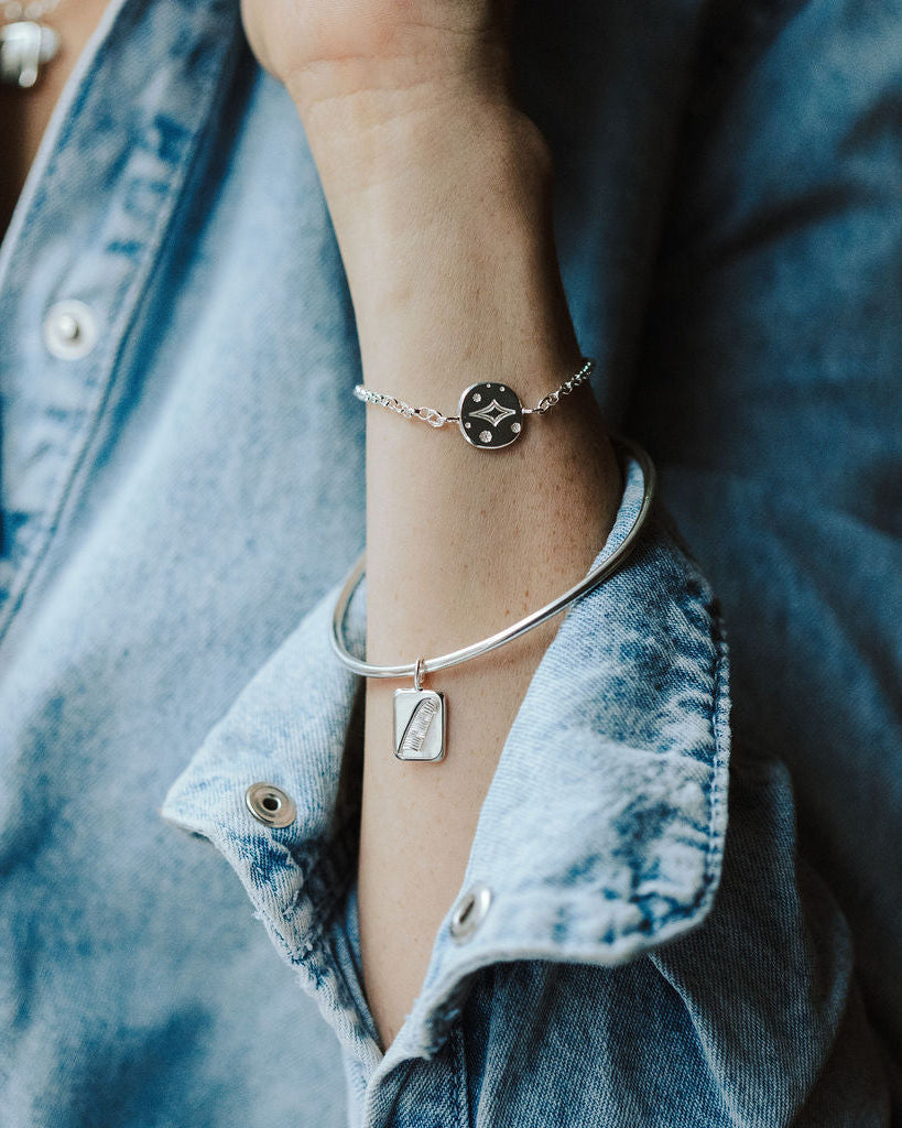 Woman in a denim shirt wearing a bangle featuring a feather charm and a bracelet featuring a star charm on her wrist.