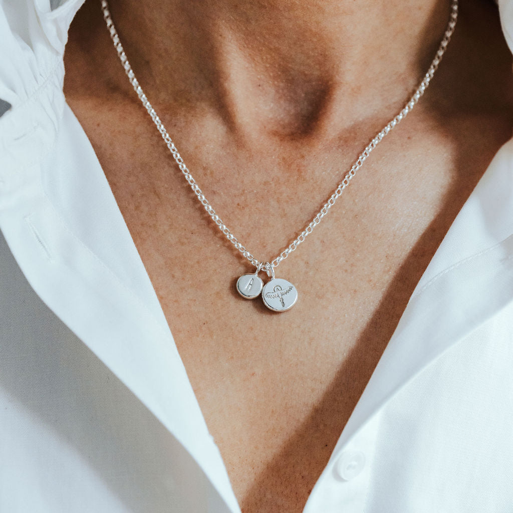 Silver necklace with round phoenix and initial pendants on a person wearing a white shirt.