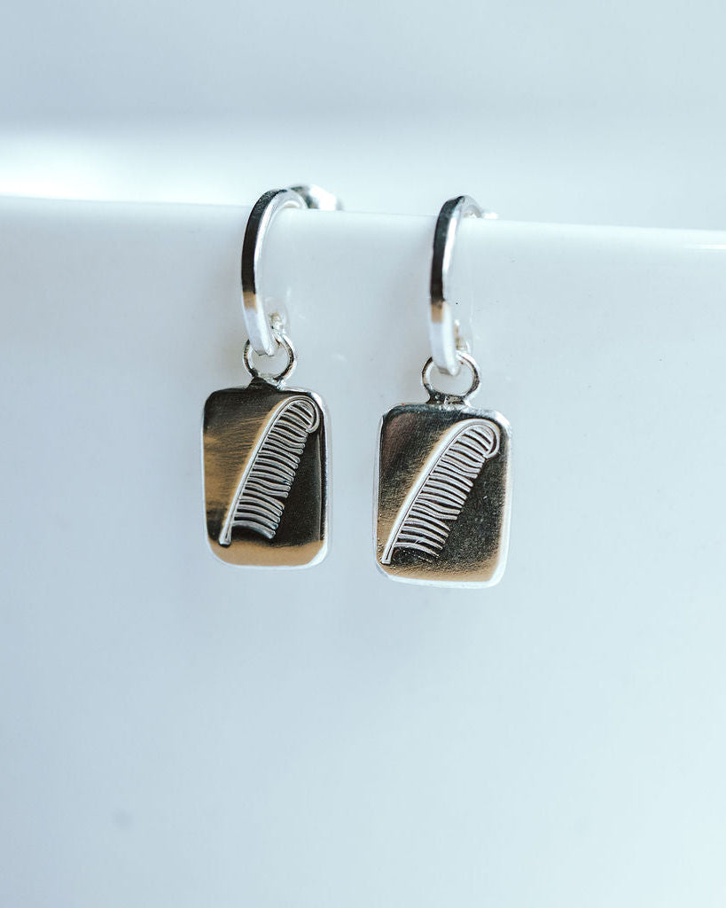 Sterling silver hoop earrings resting on a white bowl, featuring a charm with a feather design.