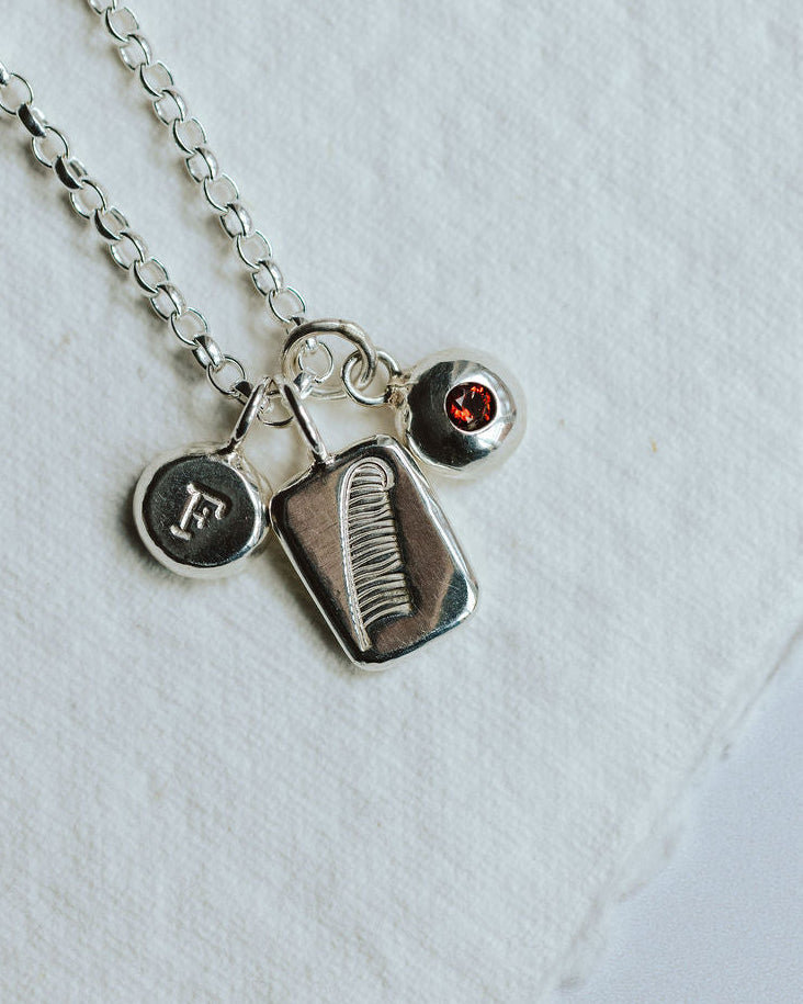 Charm necklace on a white background featuring a feather, an initial and a birthstone.