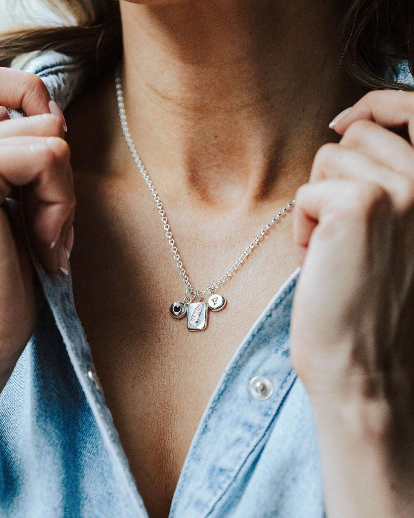 Woman wearing a denim shirt and a charm necklace featuring a feather charm, initial charm and birthstone charm.