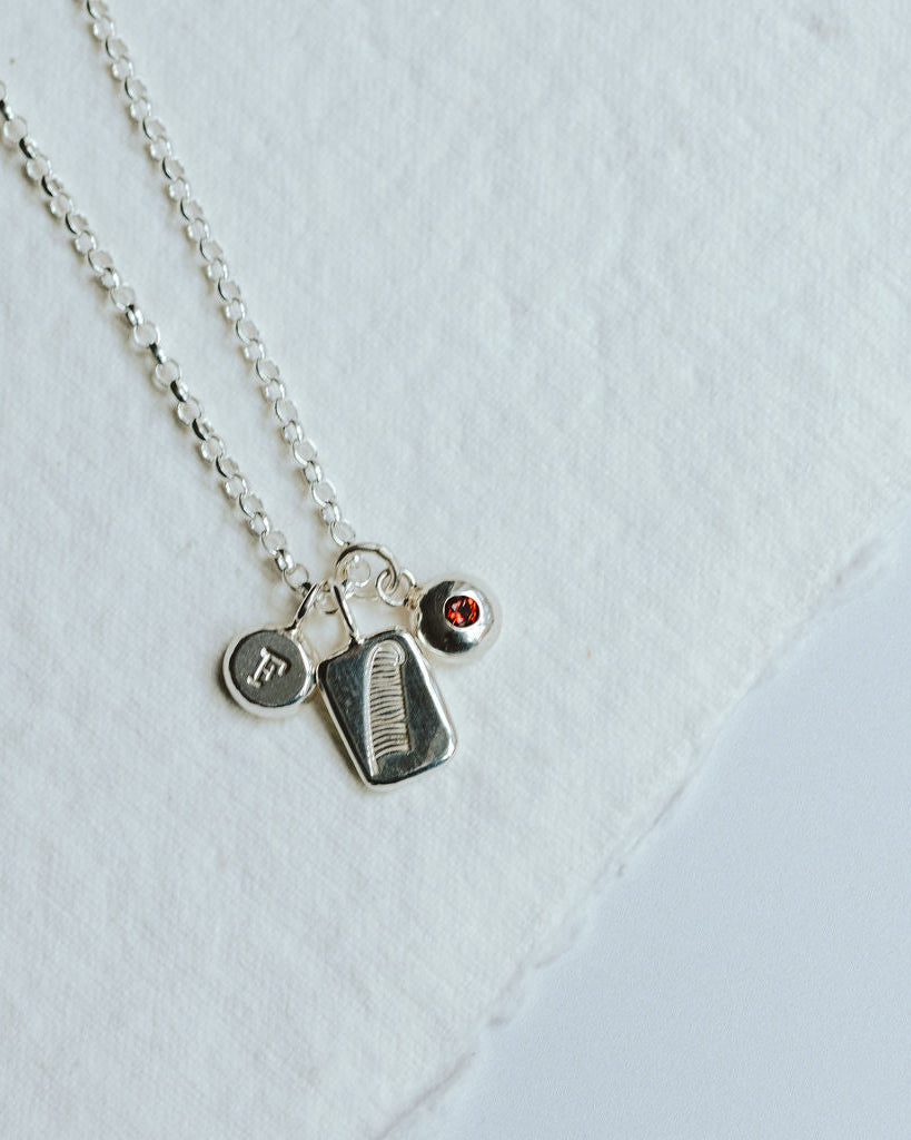 Necklace with three charms on a white background, featuring a feather design, and initial and a birthstone charm.