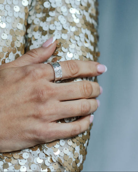 Close-up of a hand wearing a bark textured ring on a sequinned surface with a blurred background