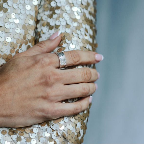 Close-up of a hand wearing a bark textured ring on a sequinned surface with a blurred background