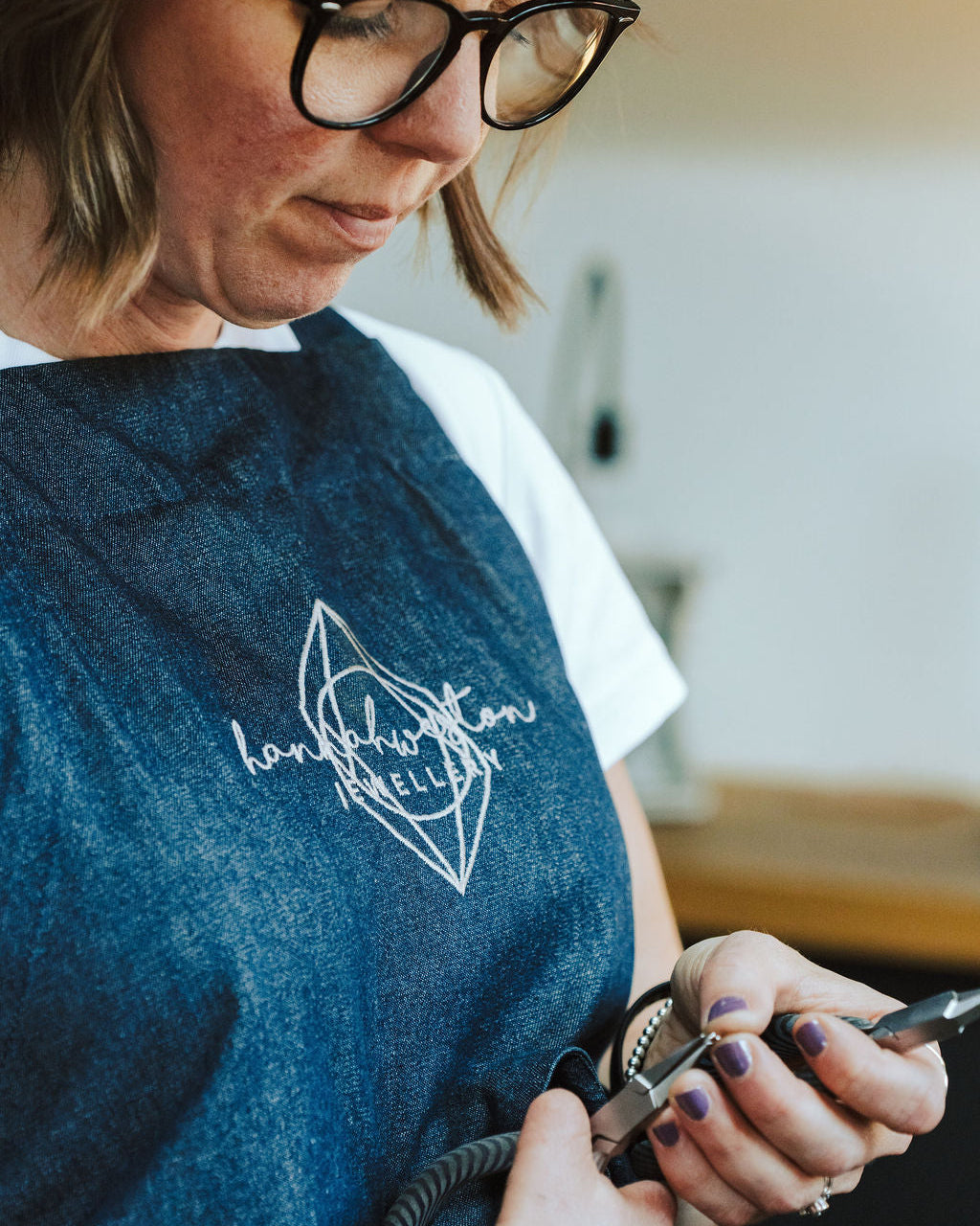 A close-up lifestyle shot of a woman wearing a blue denim apron with a white embroidered logo that reads 'hampshire Jewellery'. She is wearing black-rimmed glasses and a white t-shirt, holding a metal tool in a workshop setting.