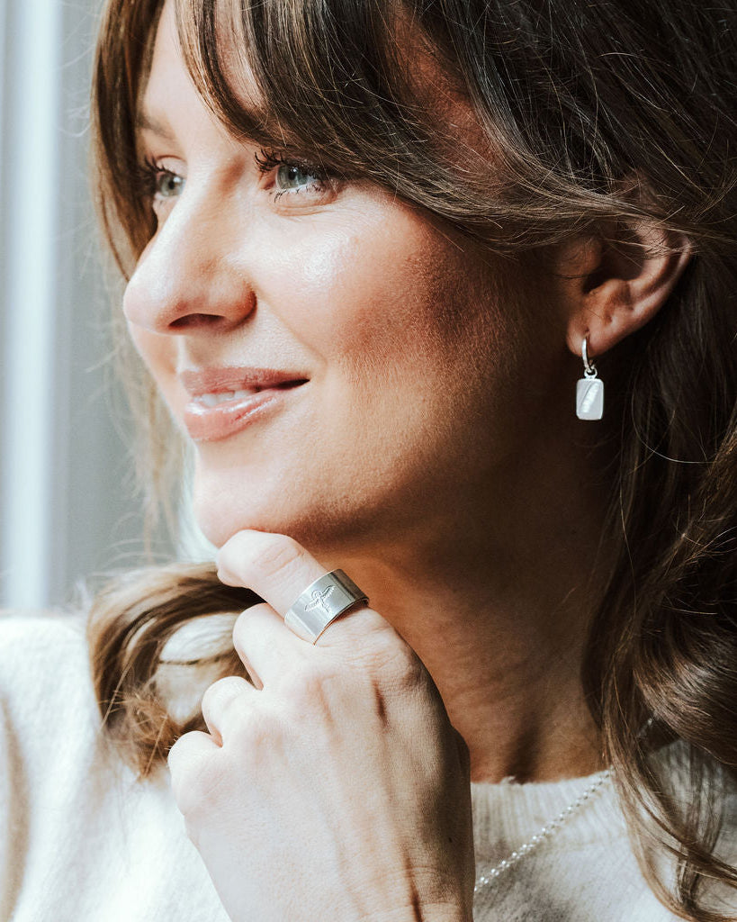 Woman with dark hair and a white knitted t-shirt wearing silver feather charm hoop earrings and a ring stamped with a phoenix design.