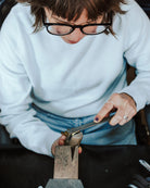 A close-up lifestyle shot of a jeweler wearing black-rimmed glasses and a white sweater, using calipers to measure a piece of jewelry held in a wooden stand on a workbench.