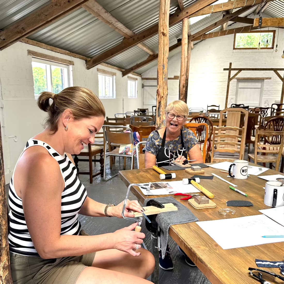 Two women sitting at a wooden table in a rustic indoor setting, at a jewellery workshop