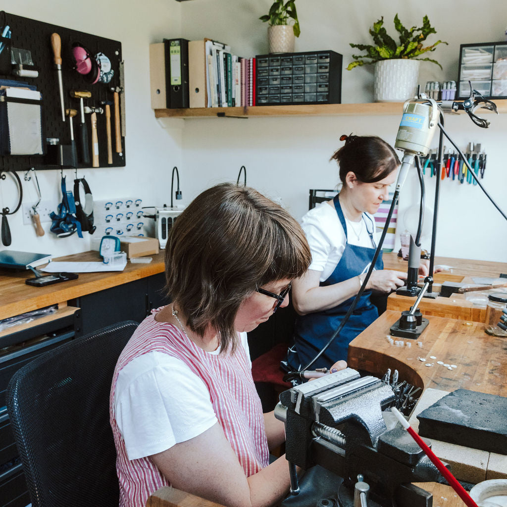 Two women working at a wooden craft desk in a workshop, one in the foreground using a black bench grinder and another in the background at her own station. The room has white walls, wooden shelves with plants and binders, and a dark ceiling.