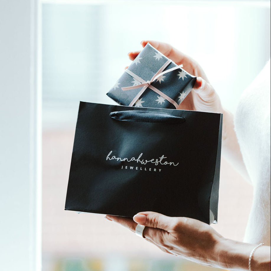 Woman holding a navy jewellery gift bag near a white framed window, lifting out a gift wrapped jewellery box.