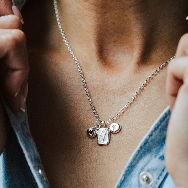 Woman in a denim shirt wearing a necklace featuring three charms, one birthstone, one feather and one initial.