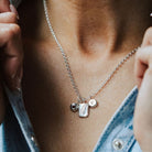 Woman in a denim shirt wearing a necklace featuring three charms, one birthstone, one feather and one initial.