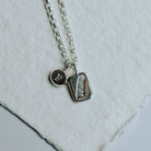 Necklace with two charms, one featuring a pendant with a feather design and the other with the letter F resting on a white background.