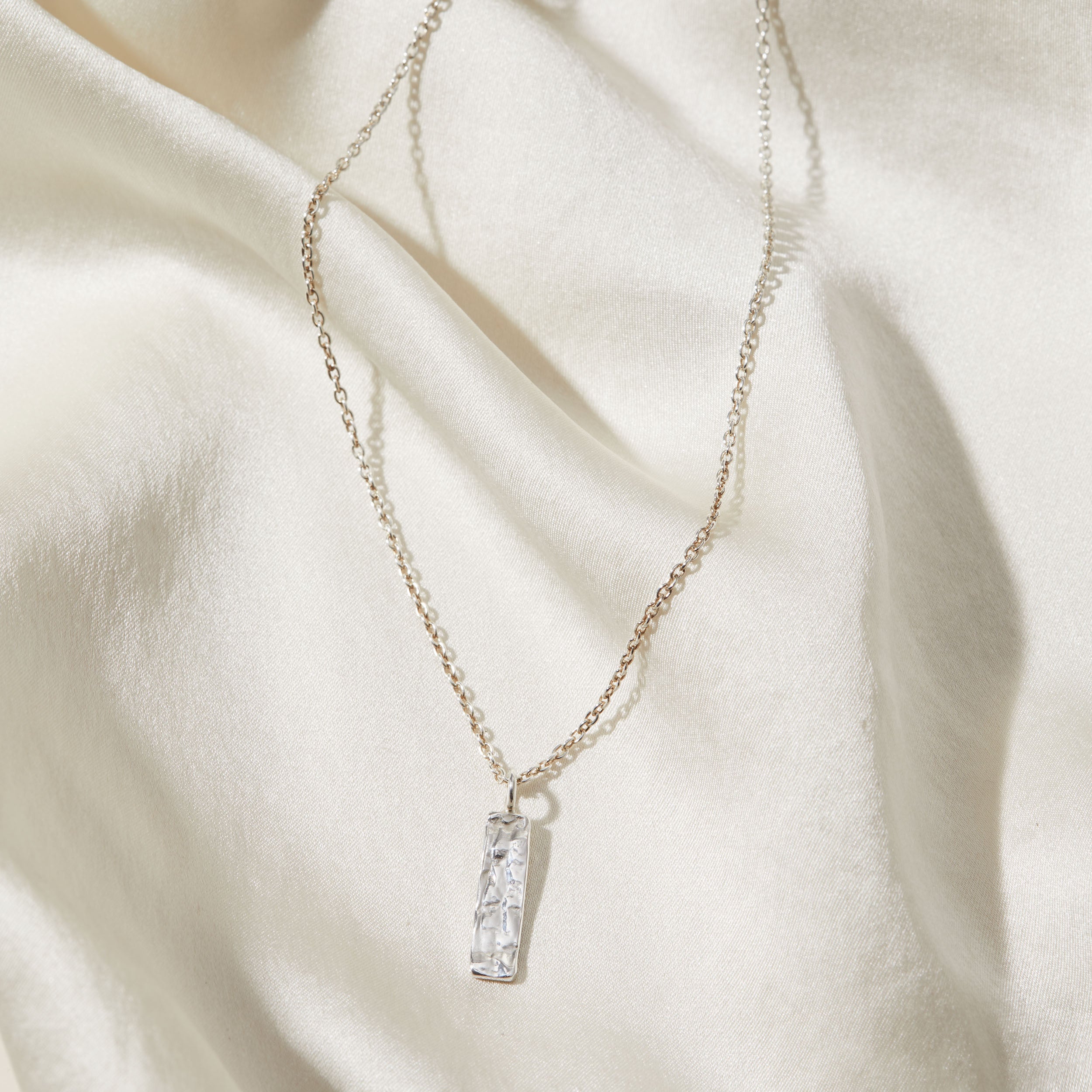 Silver necklace with a rectangular bark textured pendant on a beige fabric background