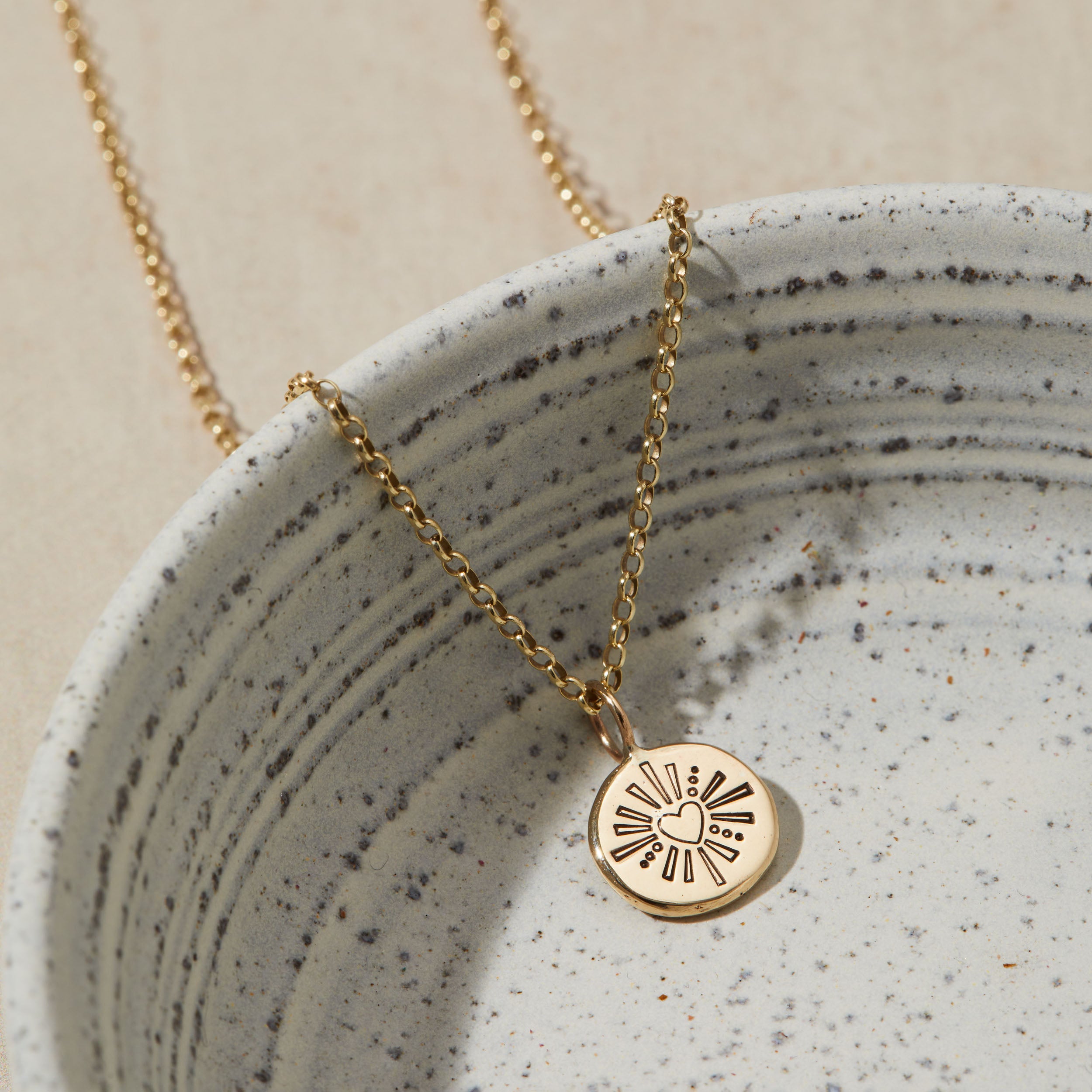 Gold necklace with a heart and sun design pendant on a speckled ceramic dish.