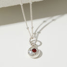 Silver necklace with a round pendant featuring a red gemstone on a white surface.