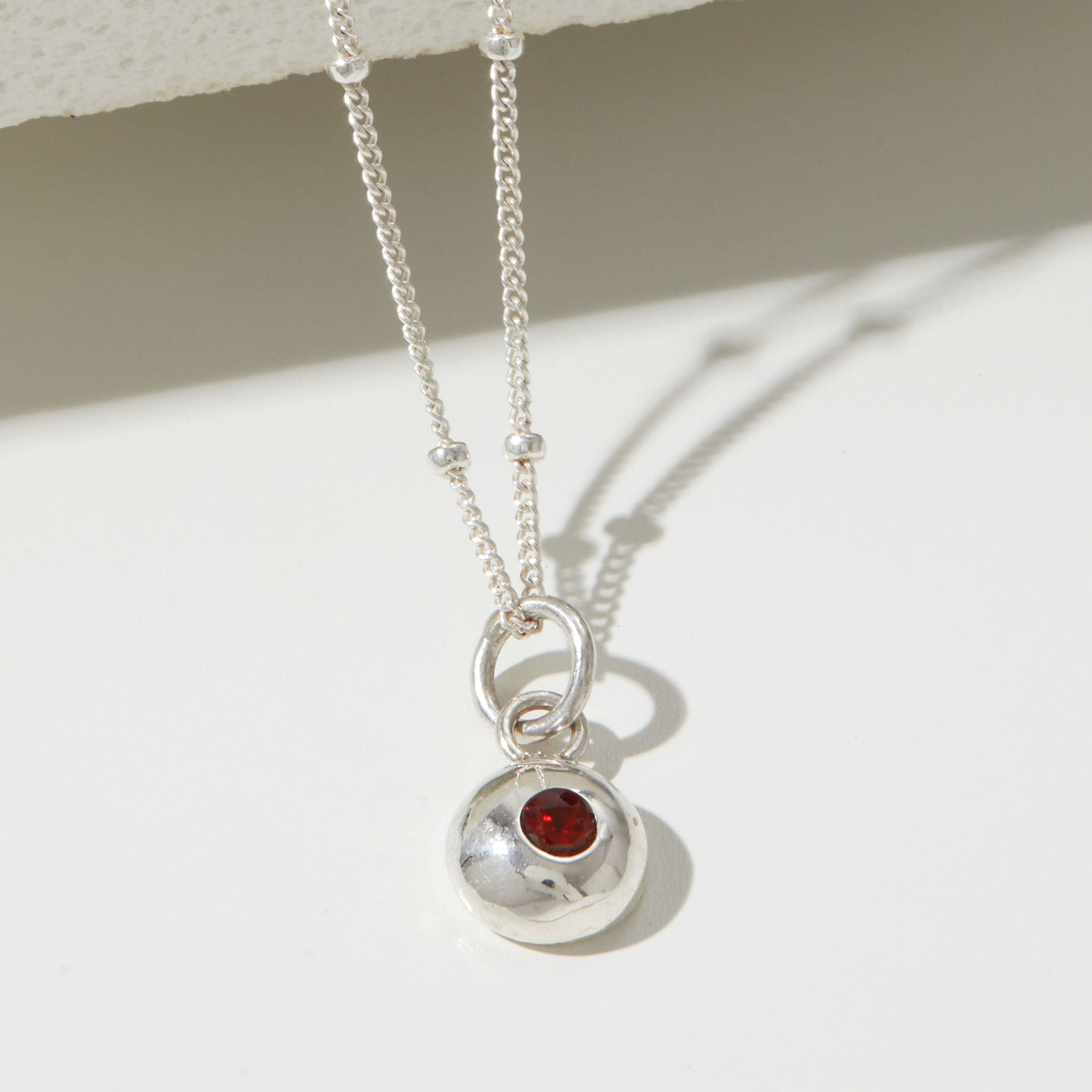 Silver necklace with a round pendant featuring a red gemstone on a white surface.