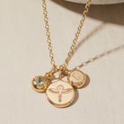 Gold necklace with birthstone, phoenix and initial charms on a beige background