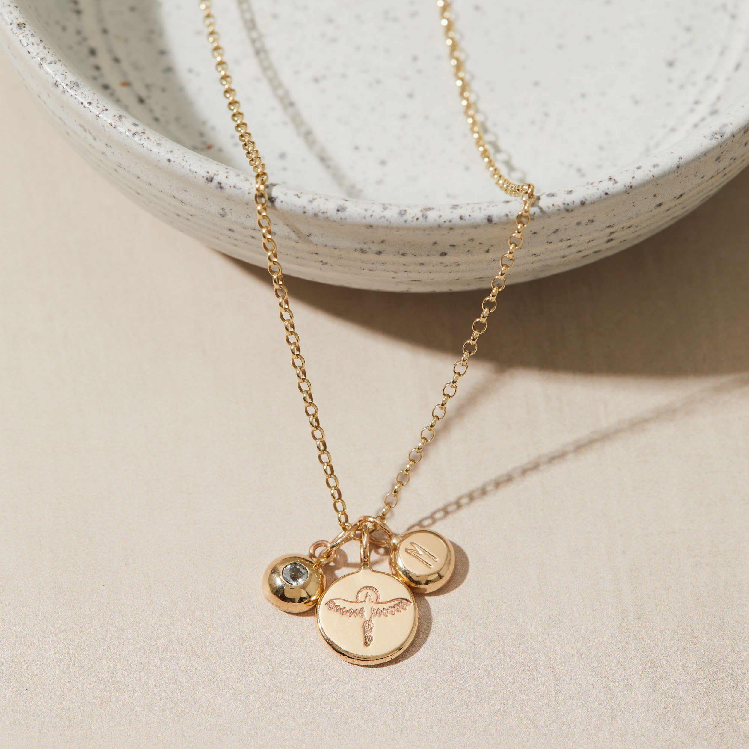 Gold necklace with initial, birthstone and phoenix pendants on a beige surface with a white textured bowl in the background.