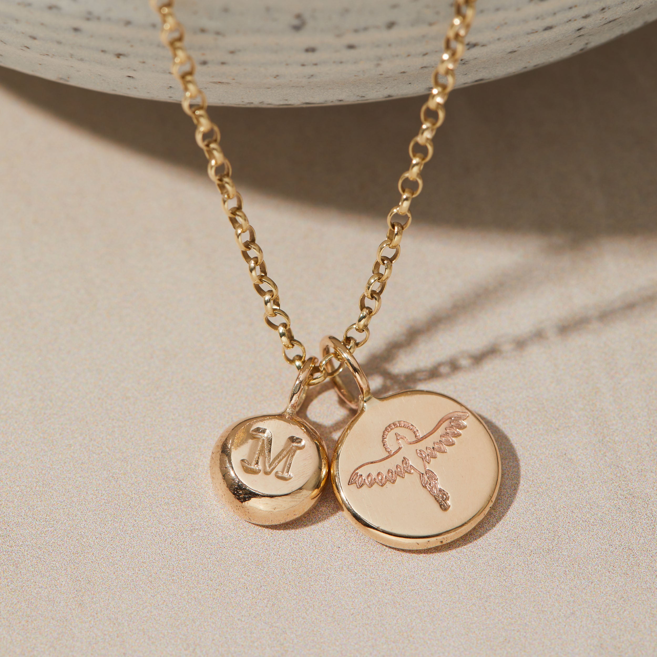 Gold necklace with initial and phoenix pendants on a beige background