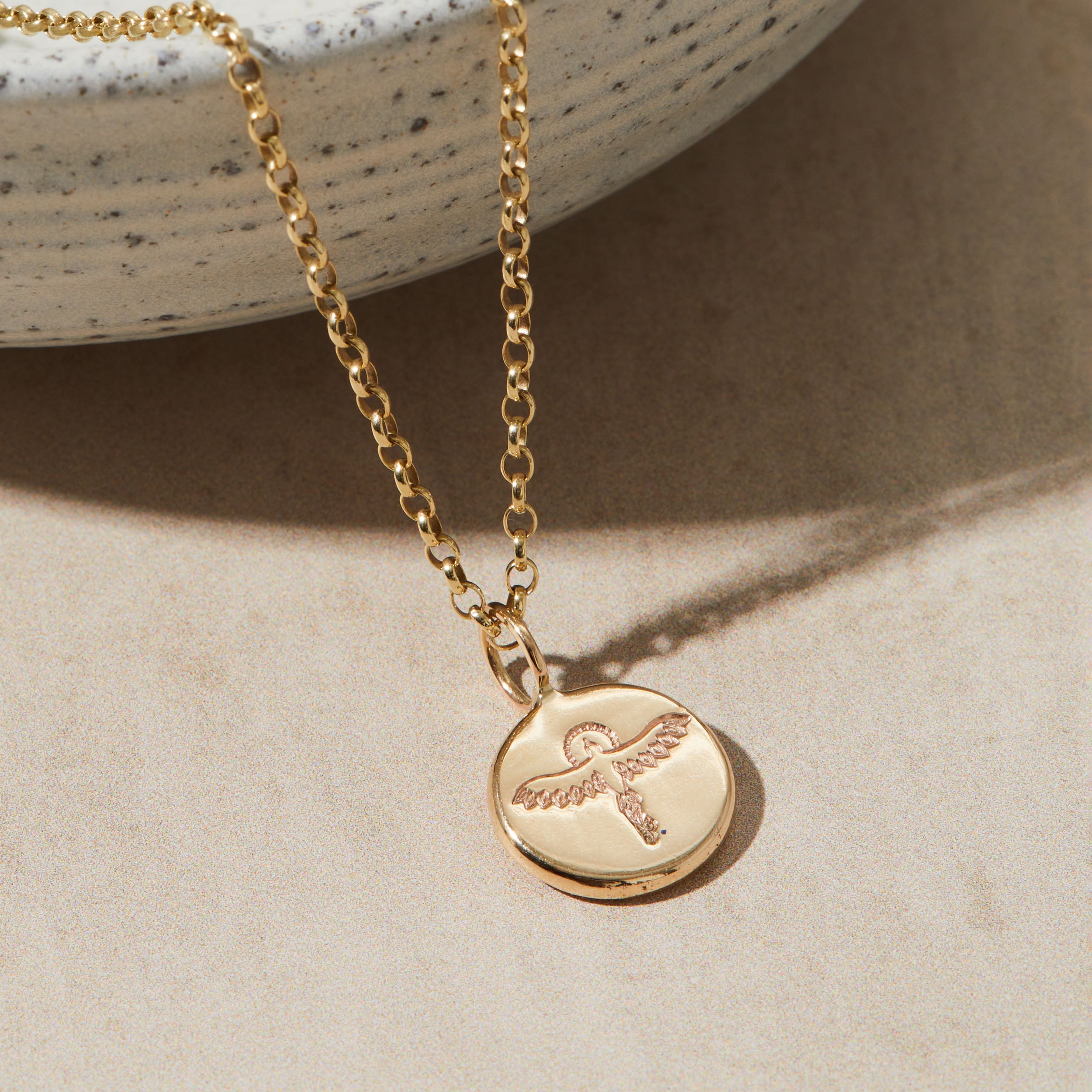 Gold necklace with a round pendant featuring an stamped phoenix design on a beige surface.