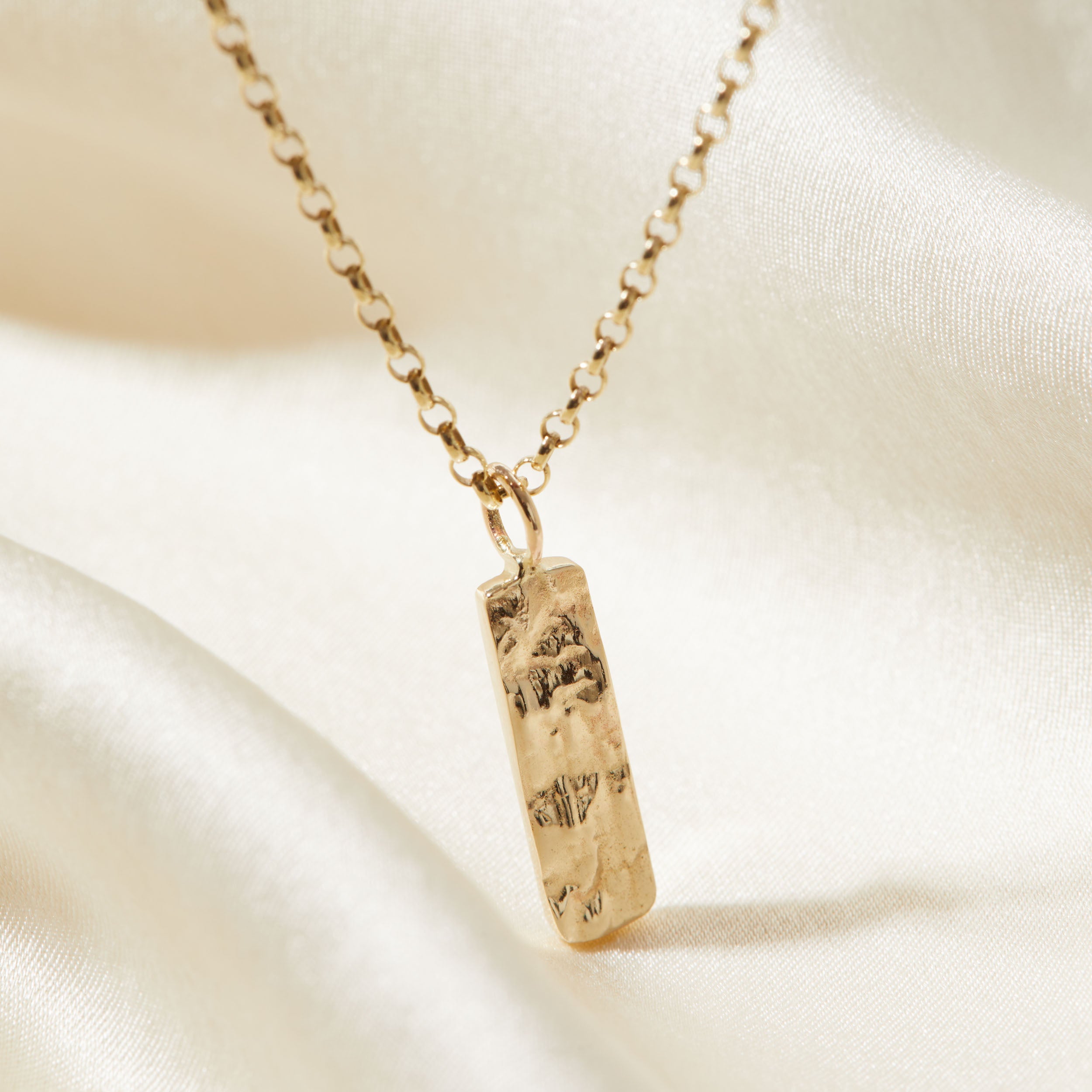 Gold necklace with a rectangular tree textured pendant on a beige fabric background