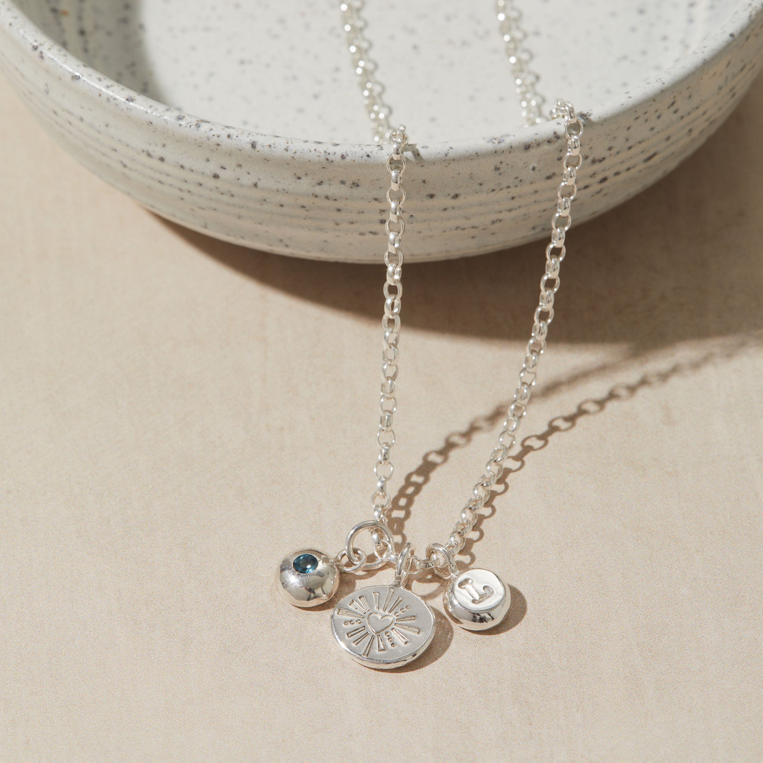 Silver necklace with heart charm, initial charm and birthstone charm on neutral surface.