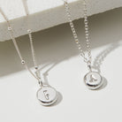 Two silver necklaces with round pendants displaying letters 'F' and 'A' on a light background.