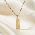 Gold necklace with a rectangular bark textured pendant on a beige fabric background