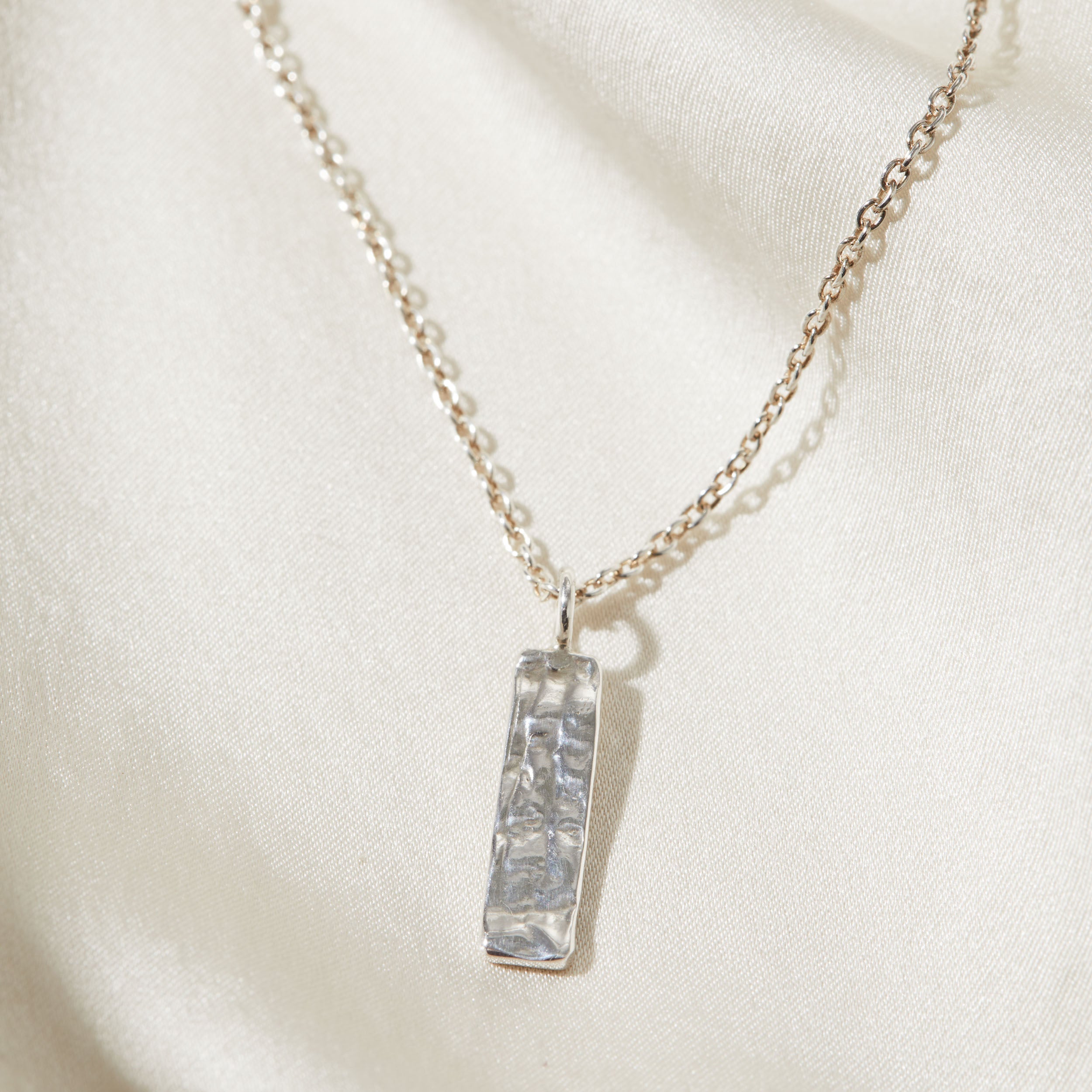 Silver necklace with a rectangular bark textured pendant on a white fabric background