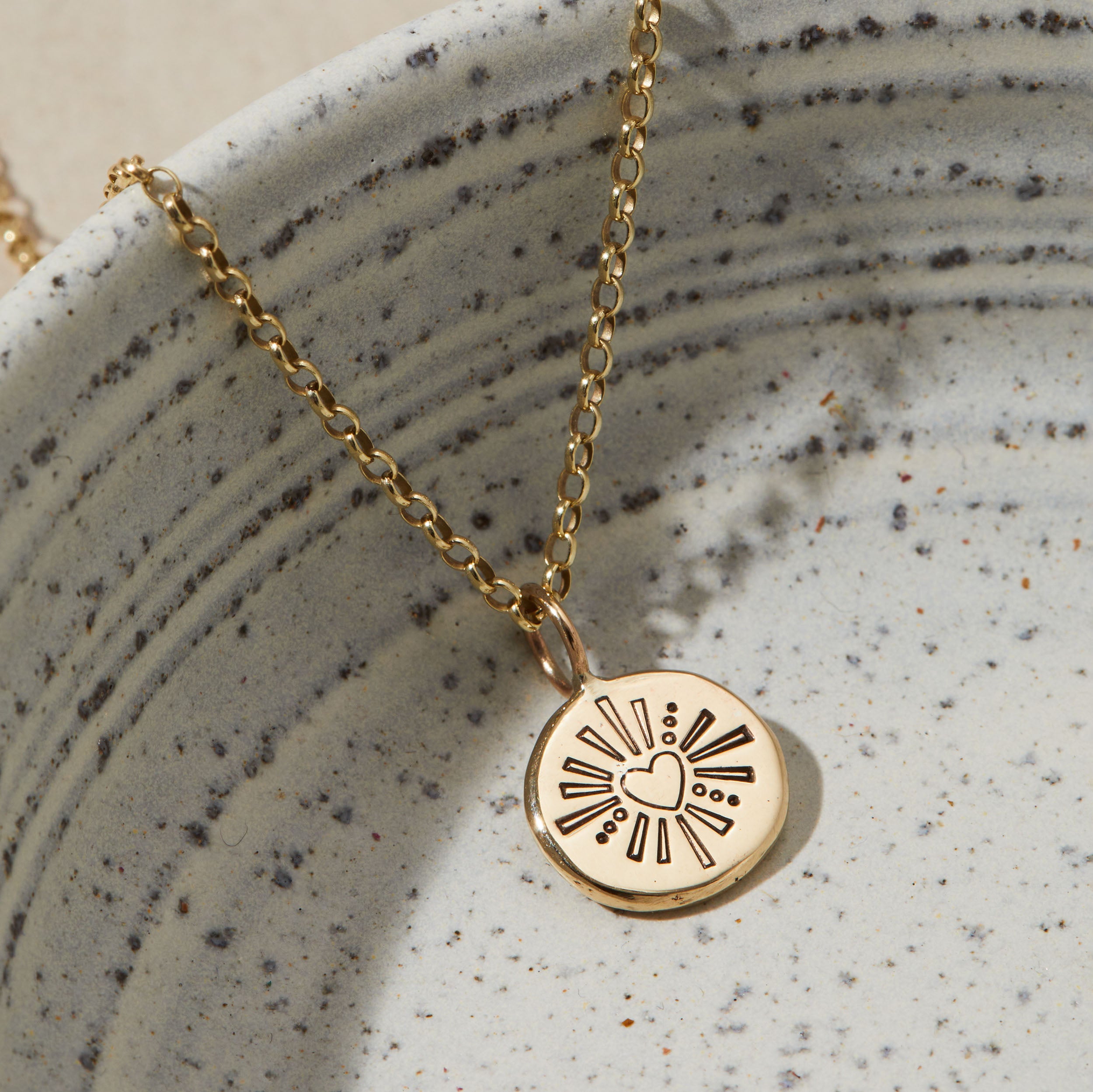 Gold necklace with a circular pendant featuring a heart design on a textured ceramic surface.