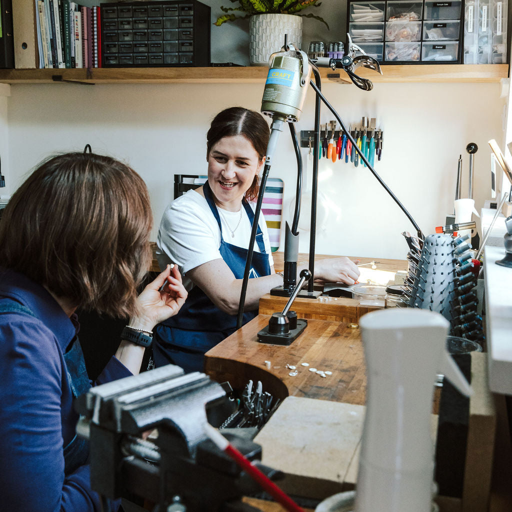 A lifestyle scene in a jewelry workshop showing a crafter smiling while working at a wooden craft table with a jeweler's loupe and tools, with another person seated in the foreground.